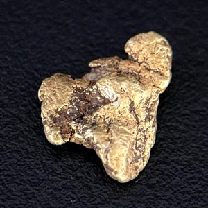 May include: A raw, naturally formed gold nugget with an irregular, textured surface. The piece displays a metallic yellow luster with deep, dark brown mineral inclusions embedded within its crevices, showcasing a jagged and organic shape.