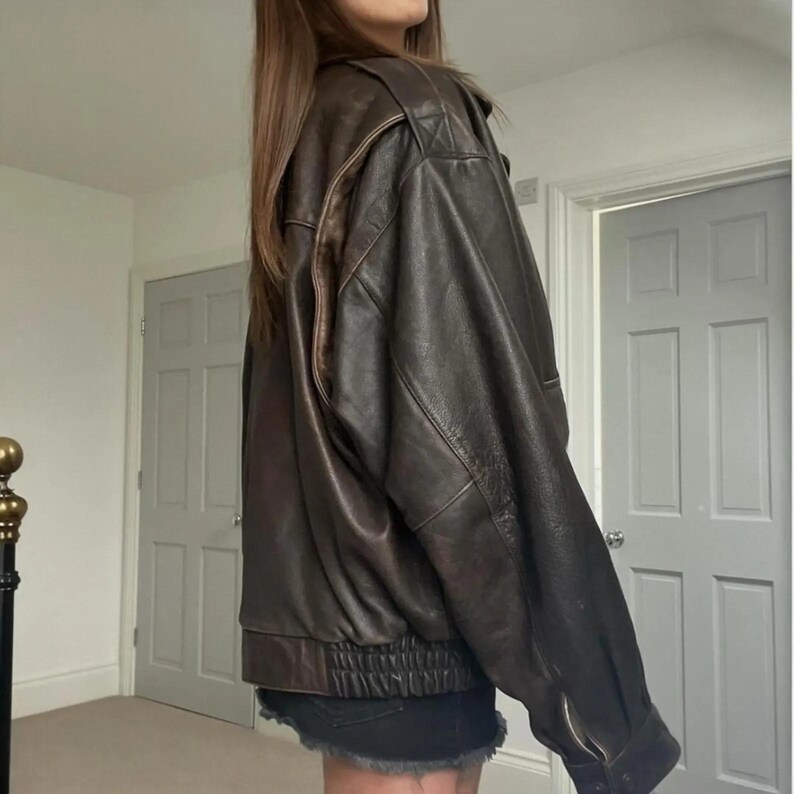 May include: A brown leather jacket with a bomber design. It has a ribbed hem and cuffs, a zip-up front, and a classic collar. The leather has a distressed appearance, giving it a vintage feel. The jacket is a medium size.