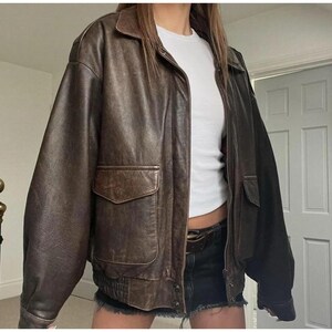May include: A brown leather jacket with a collared neckline, two front pockets, and a zip closure. The jacket has a ribbed hem and cuffs. The leather has a worn appearance, giving it a vintage aesthetic.