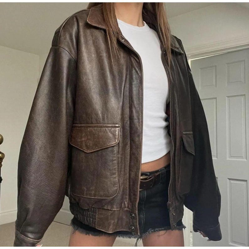May include: A brown leather jacket with a collared neckline, two front pockets, and a zip closure. The jacket has a ribbed hem and cuffs. The leather has a worn appearance, giving it a vintage aesthetic.