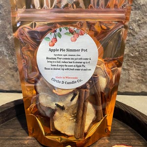 May include: A clear, metallic-gold pouch containing apple slices, cinnamon sticks, and cloves. The label reads "Apple Pie Simmer Pot" with instructions and ingredients. The product is from Circle D Candle Co., made in Wisconsin.