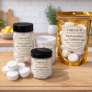 May include: Several containers of dishwasher tablets are arranged on a wooden cutting board. The tablets are white and cylindrical. The containers include jars with black lids and a gold foil bag. Text on the labels reads "CIRCLE D Dishwasher Tablets 100% All Natural."