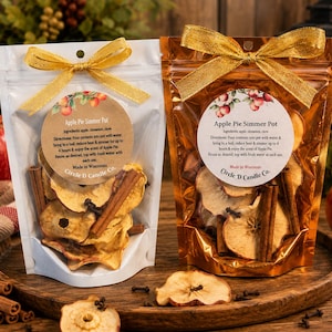 May include: Two stand-up pouches containing dried apple slices, cinnamon sticks, and cloves. The left pouch is white, the right is metallic copper, both topped with gold bows. Each features a circular label reading Apple Pie Simmer Pot.