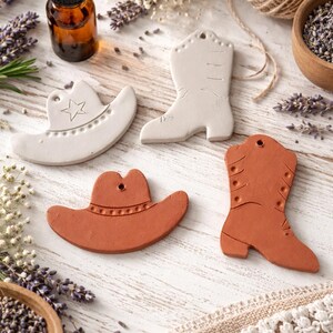 May include: Four ceramic cowboy-themed ornaments: a white cowboy hat with a star, a white cowboy boot, and a terracotta-colored cowboy hat and boot. The ornaments are arranged on a white wooden surface with lavender and essential oil.