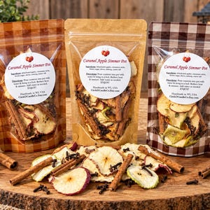 May include: Three stand-up pouches of Caramel Apple Simmer Pot mix featuring dried apple slices, cinnamon sticks, and cloves. Each pouch has a white circular label with instructions and ingredients. Loose ingredients are scattered in the foreground.