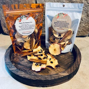 May include: Two bags of Apple Pie Simmer Pot with dried apple slices, cinnamon sticks, and cloves. One bag is clear, the other is orange. The bags are on a dark wooden tray. The text "Apple Pie Simmer Pot" is visible.