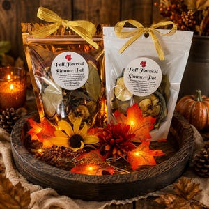May include: Two clear pouches labeled Fall Harvest Simmer Pot by Circle Candle Co. contain dried fruit and spices. Each pouch is topped with a gold ribbon. The pouches sit on a dark wooden tray decorated with artificial autumn leaves and flowers.