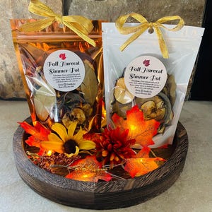 May include: Two Fall Harvest Simmer Pot pouches with gold bows, displayed in a wooden tray with fall foliage and fairy lights. The pouches contain dried botanicals. The text on the pouches reads "Fall Harvest Simmer Pot".