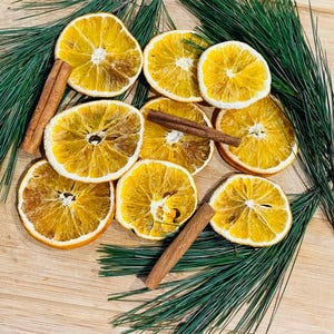 May include: Overhead shot of dried orange slices, cinnamon sticks, and green pine needles arranged on a wooden surface. The orange slices are circular and golden brown. The cinnamon sticks are brown and cylindrical. The pine needles are long and green.