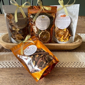 May include: Four clear pouches filled with dried fruit and spices, tied with gold ribbons. The pouches are labeled "Apple Pie Simmer Pot", "Orange Simmer Pot", "Pumpkin Spice Simmer", and "Fall Harvest Simmer Pot". The pouches are displayed in a wooden bowl.