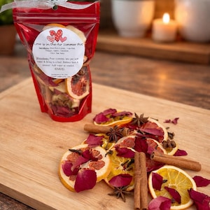 May include: A red bag of potpourri with a silver ribbon, labeled "Love is in the Air Simmer Pot." The bag contains dried orange slices, rose petals, and cinnamon sticks. Some ingredients are scattered on a wooden cutting board.