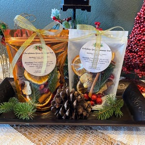 May include: Two clear pouches of potpourri, one orange and one white, with gold ribbon bows. The orange pouch is labeled "Christmas Spice Simmer Pot" and the white pouch is labeled "Holiday Spice Simmer Pot". The pouches contain dried fruit, spices, and greenery.