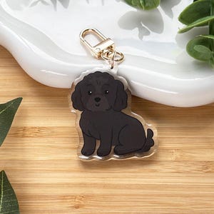 May include: A clear acrylic keychain featuring a black dog illustration with a gold-colored clasp. The dog is sitting, with a fluffy coat and a friendly expression. The keychain is on a white, wavy dish, with green leaves on a wooden surface.