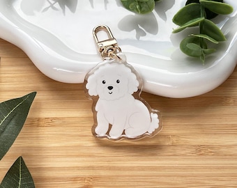 May include: A clear acrylic keychain featuring a white Bichon Frise dog illustration with a gold-colored clasp. The dog is sitting with its front paws forward. The keychain is on a white ceramic dish.