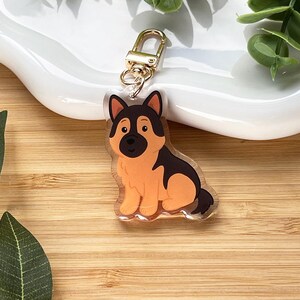 May include: A clear acrylic keychain featuring a cartoon German Shepherd dog with tan and black markings. The keychain has a gold-colored clasp and is sitting on a white surface. The background is a wooden surface with green leaves.