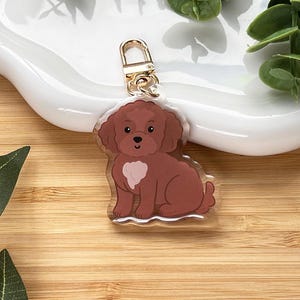 May include: A brown dog-shaped keychain with a gold-colored clasp. The dog has a white heart-shaped marking on its chest and is sitting down. The keychain is on a white, wavy-edged plate, with green leaves in the background.