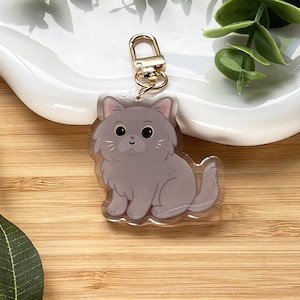May include: A gray cat-shaped keychain with a gold-colored clasp. The cat has a fluffy appearance with a smiling face and is sitting down. The keychain is on a wooden surface.