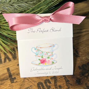 May include: A white favor box with a pink satin ribbon bow. The box features the text "The Perfect Blend" and a watercolor illustration of stacked teacups with floral accents. The names "Gabrielle and Joseph" and the date "January 1, 2026" are also printed.