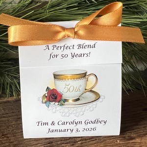 May include: A white gift box tied with a gold ribbon bow. The box features a teacup illustration with gold trim and a red rose. Text reads "A Perfect Blend for 50 Years!" and includes names and a date.