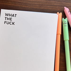 May include: A white notepad with the text "WHAT THE FUCK" in black lettering.  Five colorful pens with cat-shaped tops are arranged to the right of the notepad. The pens are orange, green, pink, purple, and tan.