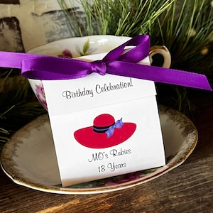 May include: A white card with a red hat illustration and the text "Birthday Celebration!" is tied to a teacup with a purple ribbon. The card also reads "MO's Rubies 18 Years". The teacup sits on a saucer with a gold rim.