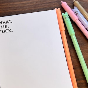 May include: A white notepad with the text "WHAT. THE. FUCK." is next to several colorful pens. The pens are orange, green, pink, purple, and gold. The pens have cat-shaped tops. The notepad and pens are on a wooden surface.
