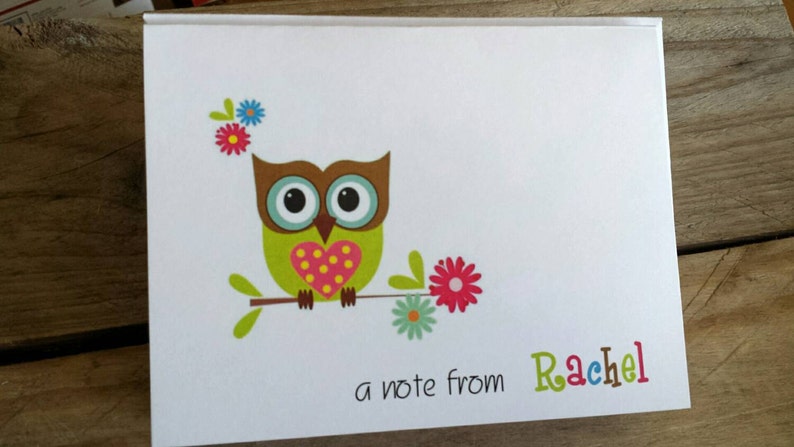 May include: A white notebook with a colorful owl illustration. The owl has a green body, brown head, and a pink heart. The text "a note from Rachel" is written in colorful letters.