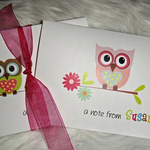 May include: Two white note card sets tied with a sheer burgundy ribbon. Each card features a colorful cartoon owl perched on a branch with flowers. One owl is green and brown, the other is pink. Text reads "a note from Susan."