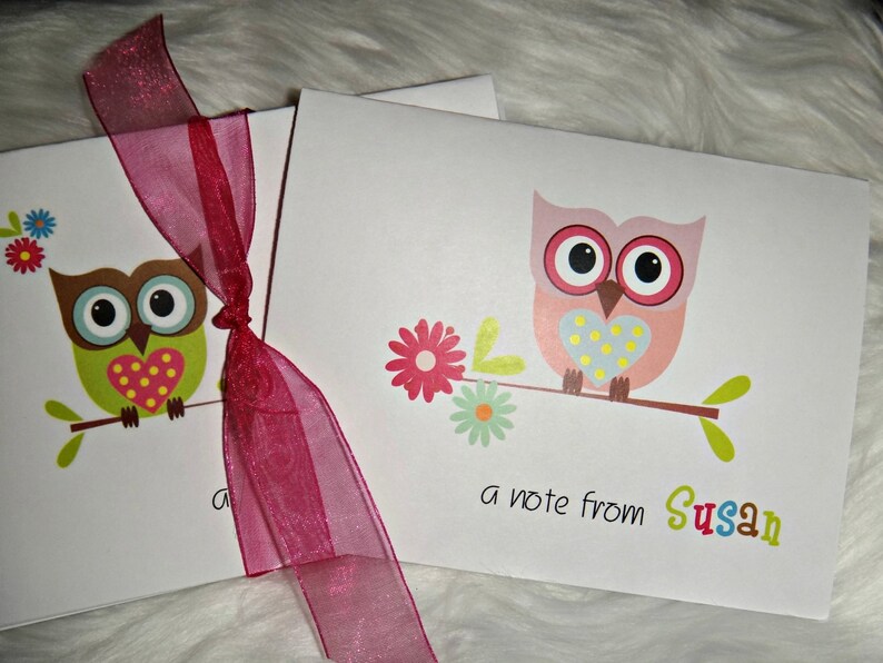 May include: Two white note card sets tied with a sheer burgundy ribbon. Each card features a colorful cartoon owl perched on a branch with flowers. One owl is green and brown, the other is pink. Text reads "a note from Susan."
