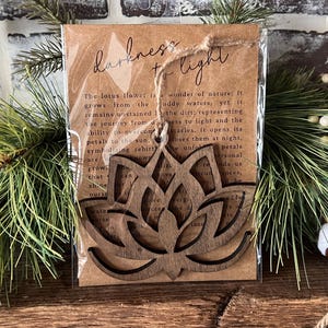 May include: A wooden lotus flower ornament with the words "Darkness to Light" on a brown card. The card has text about the lotus flower's symbolism. The ornament is attached with twine and is displayed with evergreen branches.