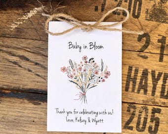 May include: A white card with the text "Baby in Bloom" and a floral illustration. The card is tied with twine and includes the text "Thank you for celebrating with us! Love, Kelsey & Wyatt."