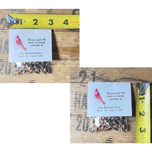 two small packages of birdseed placed on a wooden surface. The packages are labeled with a message about the memory of a loved one, likely a bird. The image also includes a yellow measuring tape, which is placed on the wooden surface.