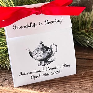 May include: A white gift box with a red bow and the text "Friendship is Brewing!". The box features a black and white illustration of a teapot and the text "International Reunion Day April 15th, 2023".