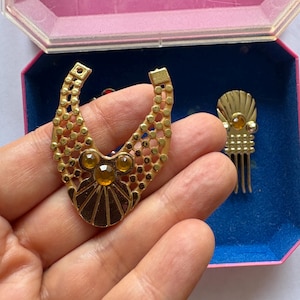 Peut inclure: Ensemble de bijoux vintage dor&eacute;s, comprenant un grand pendentif et une petite pi&egrave;ce en forme de peigne. Le pendentif pr&eacute;sente un design g&eacute;om&eacute;trique avec des accents de couleur ambre. Les bijoux sont pr&eacute;sent&eacute;s dans une bo&icirc;te rose et bleue.