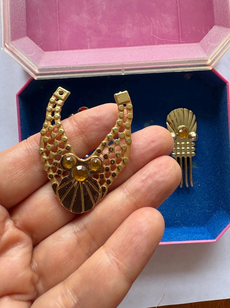 Peut inclure: Ensemble de bijoux vintage dor&eacute;s, comprenant un grand pendentif et une petite pi&egrave;ce en forme de peigne. Le pendentif pr&eacute;sente un design g&eacute;om&eacute;trique avec des accents de couleur ambre. Les bijoux sont pr&eacute;sent&eacute;s dans une bo&icirc;te rose et bleue.
