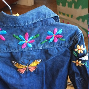 May include: A blue denim shirt with colourful embroidered flowers and a butterfly on the back.