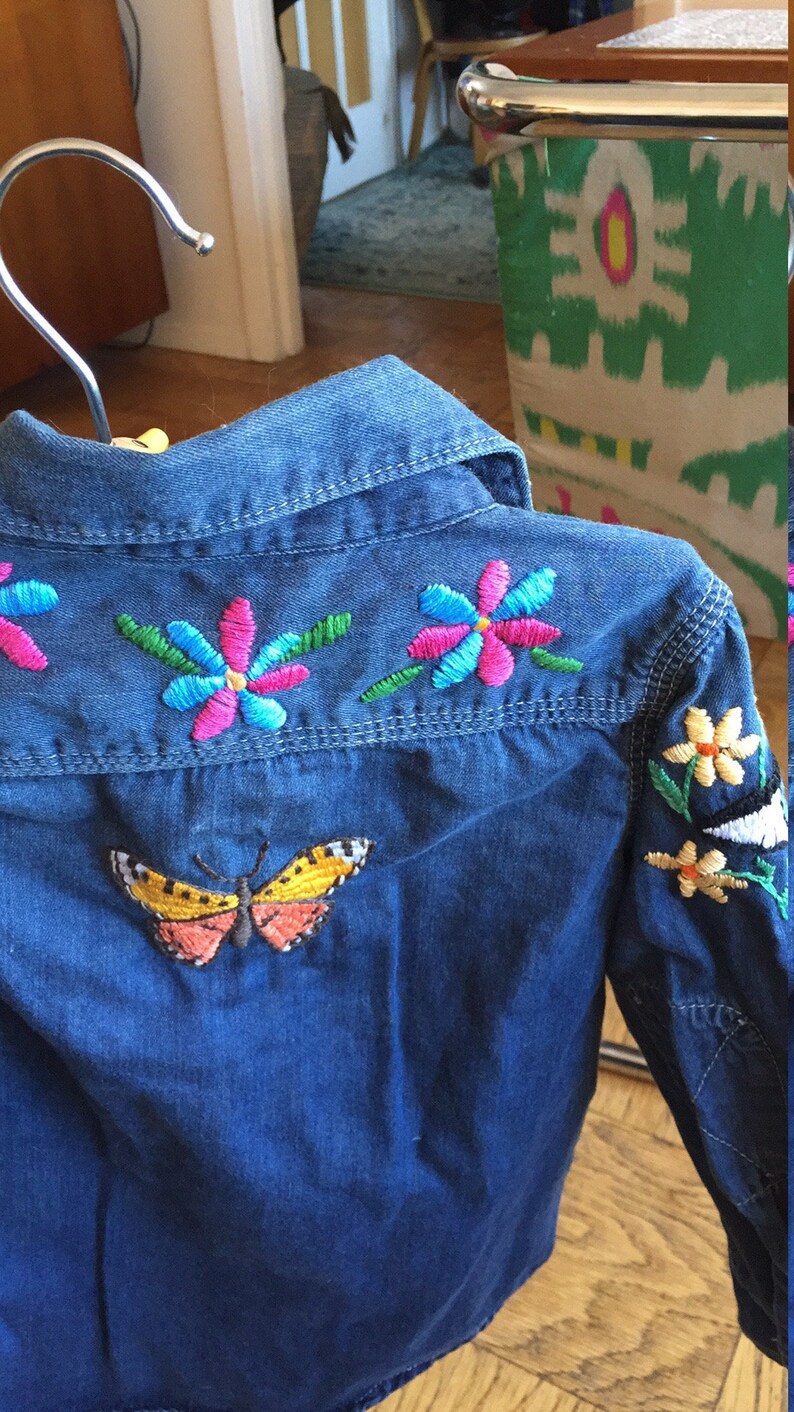 May include: A blue denim shirt with colourful embroidered flowers and a butterfly on the back.