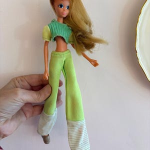 May include: A vintage doll with long blonde hair and blue eyes, dressed in a green and yellow outfit. The doll wears a cropped green top, green trousers, and brown shoes. The trousers have white and green striped cuffs.