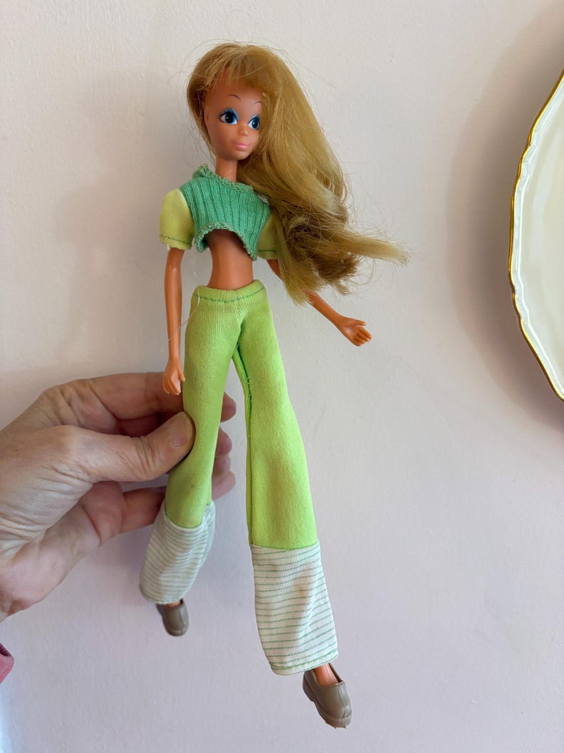 May include: A vintage doll with long blonde hair and blue eyes, dressed in a green and yellow outfit. The doll wears a cropped green top, green trousers, and brown shoes. The trousers have white and green striped cuffs.