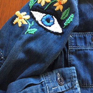 May include: A blue denim jacket with a large embroidered eye and yellow flowers on the sleeve. The jacket has a button fastening and a pocket on the front.