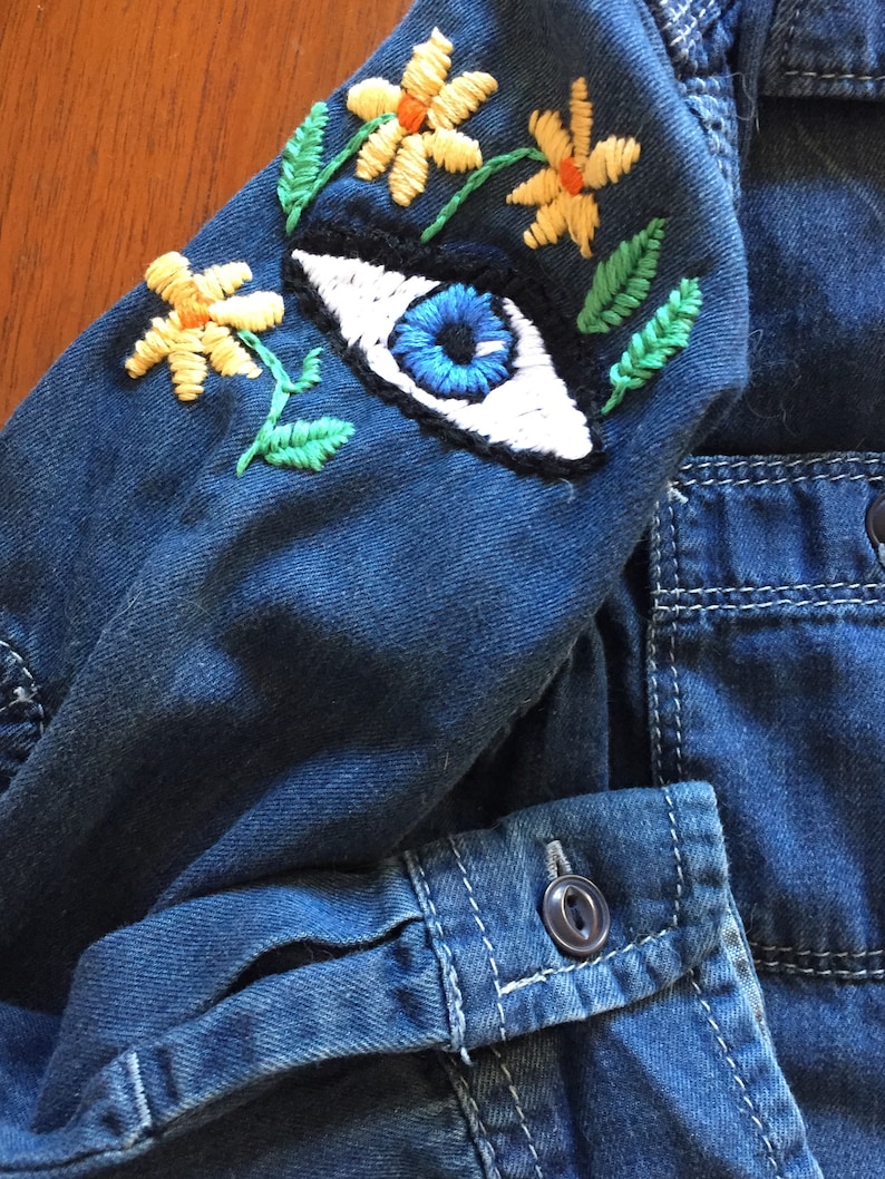 May include: A blue denim jacket with a large embroidered eye and yellow flowers on the sleeve. The jacket has a button fastening and a pocket on the front.
