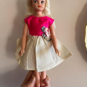 May include: A vintage doll with blonde hair and blue eyes, wearing a bright pink top and a cream-colored skirt. The doll's dress is adorned with a floral embellishment. The doll is held in a person's hand.