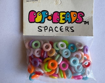 May include: A clear plastic bag filled with colorful Pop-Beads spacers. The bag has a cardboard header with the words "POP-BEADS SPACERS" in a playful font. The spacers are in various colors, including pink, green, blue, and orange.