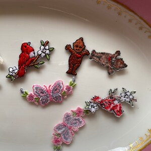 May include: Assortment of embroidered iron-on patches featuring a red bird on a branch, a butterfly, a gnome, a bear, and a red bird with white flowers.