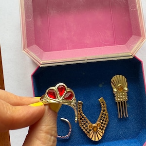 Peut inclure: Ensemble de bijoux fantaisie vintage, comprenant une bague dor&eacute;e avec des pierres rouges en forme de c&oelig;ur, un collier dor&eacute;, un peigne &agrave; cheveux et une bague argent&eacute;e, pr&eacute;sent&eacute;s dans une bo&icirc;te rose et bleue.