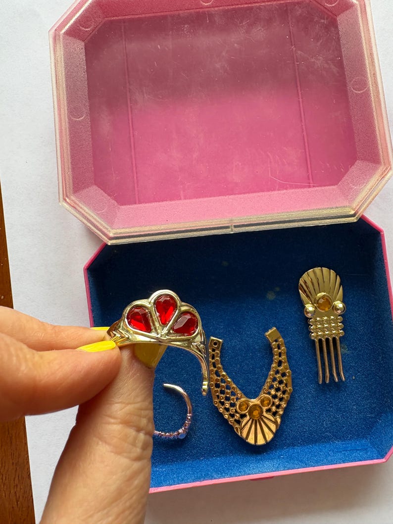 Peut inclure: Ensemble de bijoux fantaisie vintage, comprenant une bague dor&eacute;e avec des pierres rouges en forme de c&oelig;ur, un collier dor&eacute;, un peigne &agrave; cheveux et une bague argent&eacute;e, pr&eacute;sent&eacute;s dans une bo&icirc;te rose et bleue.