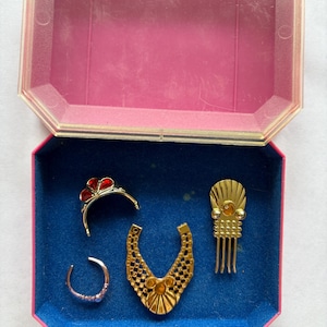 Peut inclure: Un ensemble de bijoux vintage dans une bo&icirc;te rose et bleue. L'ensemble comprend un collier dor&eacute;, un diad&egrave;me avec des accents rouges, une bague et un peigne &agrave; cheveux. La bo&icirc;te est ouverte, r&eacute;v&eacute;lant les bijoux sur une doublure en velours bleu.