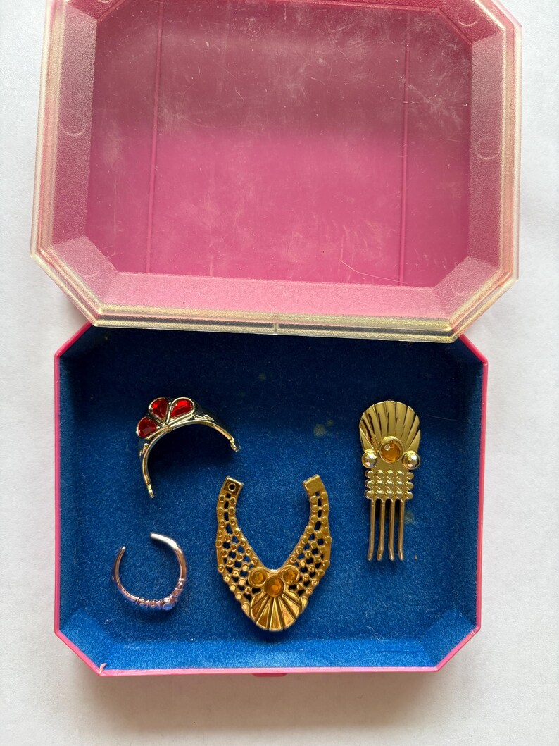 Peut inclure: Un ensemble de bijoux vintage dans une bo&icirc;te rose et bleue. L'ensemble comprend un collier dor&eacute;, un diad&egrave;me avec des accents rouges, une bague et un peigne &agrave; cheveux. La bo&icirc;te est ouverte, r&eacute;v&eacute;lant les bijoux sur une doublure en velours bleu.