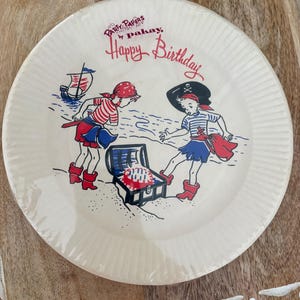 May include: Round paper plate with a pirate-themed design. The plate features two cartoon pirates, a treasure chest, and a ship. Text on the plate reads "Party Papers by Pakay" and "Happy Birthday" in red script.