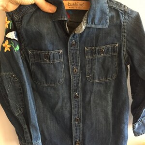 May include: A dark blue denim shirt with long sleeves and a button-up front. The shirt has two chest pockets and embroidered flowers on the left sleeve. The shirt has a label that reads "kushies baby + baba".
